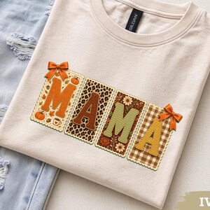 May include: An ivory crew neck t-shirt featuring the word MAMA in large block letters. Each letter has a unique pattern including pumpkins, leopard print, floral motifs, and gingham checks, accented with orange bows and scalloped edges.