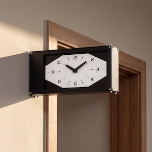 Puede incluir: Un reloj de pared rectangular con marco negro y esfera blanca octogonal. El reloj cuenta con manecillas negras para las horas y los minutos, con números negros en las posiciones de las 12, 2, 4, 6, 8 y 10, y marcas negras para las horas restantes.