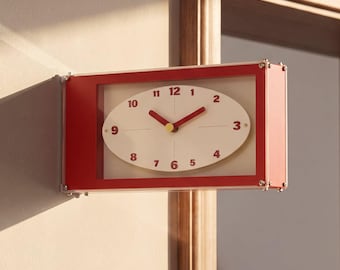 May include: A rectangular wall clock with a red frame and a white oval face. The clock features red numerals from 1 to 12, red hour and minute hands, and a yellow center point. It is mounted on a wall corner, casting a long shadow to the left.