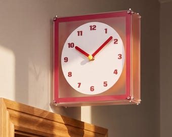 May include: A square wall clock featuring a translucent red frame and a white circular face. The clock has bold red numerals from 1 to 12, red hour and minute hands, and a small yellow center cap. The design is mounted on a flat wall surface.