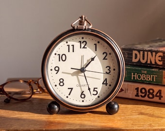 Puede incluir: Un reloj despertador analógico de cuarzo redondo con un marco de metal color bronce oscuro y una esfera de color crema. El reloj presenta números arábigos negros del 1 al 12, manecillas negras para horas, minutos y segundos, y dos pequeños pies esféricos.