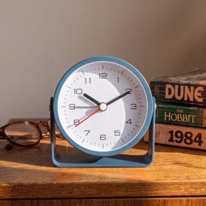 May include: A round analog desk clock with a matte blue frame and a white face. The clock features black hour and minute hands, a thin red second hand, and black numerical hour markers from 1 to 12. It is mounted on a matching blue U-shaped stand.