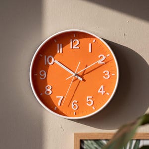 May include: A round wall clock featuring a vibrant orange face with raised white Arabic numerals and white hour, minute, and second hands. The clock is encased in a thin white frame, displaying a clear contrast between the bright orange dial and the white markers.