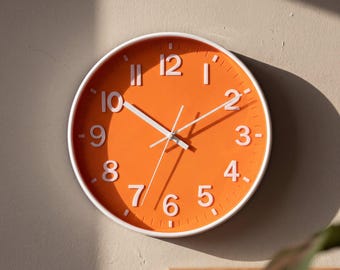 May include: A round wall clock featuring a vibrant orange face with raised white Arabic numerals and white hour, minute, and second hands. The clock is encased in a thin white frame, displaying a clear contrast between the bright orange dial and the white markers.
