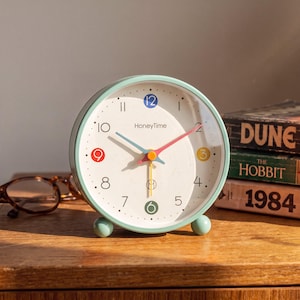 Pode incluir: Um despertador analógico redondo verde-menta com mostrador branco, apresentando o texto HoneyTime, números pretos e detalhes coloridos nas posições 12, 9, 3 e 6. O relógio possui ponteiros azul, rosa e amarelo e dois pequenos pés esféricos.