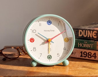 May include: A round mint green analog alarm clock with a white face, featuring the text HoneyTime, black numerals, and colorful accents on the 12, 9, 3, and 6 positions. The clock has blue, pink, and yellow hands and two small spherical feet.