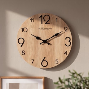 Puede incluir: Un reloj de pared redondo con una esfera de madera clara y grandes números negros. El reloj cuenta con manecillas de metal negro y el texto "est 1765 vintage clocks QUARTZ CLOCK" impreso en negro debajo de la posición de las doce.