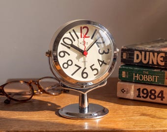 Puede incluir: Un reloj de escritorio redondo de metal plateado con esfera color crema, números negros y un número doce en rojo. El reloj cuenta con manecillas negras para horas y minutos, un segundero rojo y la palabra quartz impresa bajo el centro.