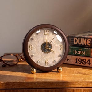 May include: A round table clock with a dark wood frame and a cream-coloured, marbled face. It features gold-toned hands, gold numerals at the 12, 3, 6, and 9 positions, and small clear crystals marking the other hours. The clock rests on two small gold-toned ball feet.