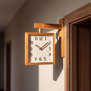 Op de afbeelding: Een vierkante wandklok met een lichthouten frame en een witte wijzerplaat. De klok heeft donkerbruine verhoogde cijfers van één tot twaalf en bijpassende donkerbruine uur- en minutenwijzers. Het is bevestigd aan een houten arm aan een deurkozijn.