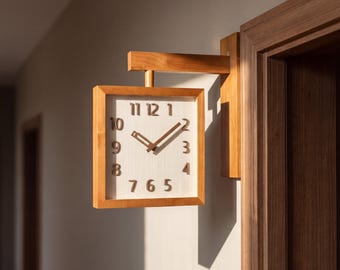 May include: A square wall-mounted clock with a light wood frame and a white face. The clock features dark brown raised numerals from one to twelve and matching dark brown hour and minute hands. It is attached to a wooden arm extending from a door frame.