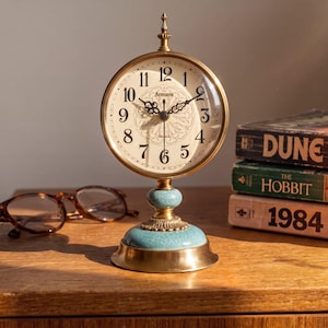 Puede incluir: Un reloj de mesa redondo de latón con esfera color crema, números arábigos negros y el texto Armans. El reloj tiene manecillas negras ornamentadas y se asienta sobre una base con un detalle de cerámica azul claro craquelada y borde de latón.