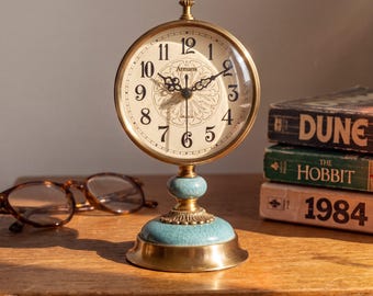 May include: A round brass table clock with a cream-colored face featuring black Arabic numerals and the text Armans. The clock has ornate black hands and sits on a pedestal base with a light blue crackle-glazed ceramic accent and brass trim.