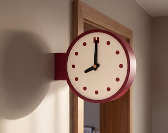 May include: A round, wall-mounted clock with a deep red frame and a cream-colored face. The clock features dark red circular hour markers, black hour and minute hands, and a small orange center point. It is mounted to the wall beside a wooden door frame.