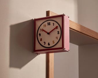 May include: A square wall clock with a pink frame and a white face featuring dark-colored numbers from 1 to 12. The clock has two red hands and is mounted on the edge of a wooden structure, casting a distinct shadow on the adjacent wall surface.
