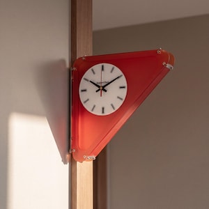 Puede incluir: Un reloj de pared para esquina con marco triangular de acrílico rojo y esfera circular blanca. La esfera muestra marcadores de hora negros, manecillas negras y el texto Gradient Clock en letras negras. Está montado en una esquina de madera.
