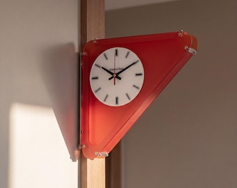 May include: A corner-mounted wall clock featuring a triangular red acrylic frame and a circular white face. The clock face displays black hour markers, black hands, and the text Gradient Clock in black lettering. It is mounted on a wooden corner.