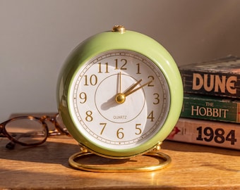 May include: A round, light green analog quartz alarm clock with a white face, gold-toned numbers, and gold-toned hands. The clock features a small gold-toned button on top and rests on a circular gold-toned metal stand base.