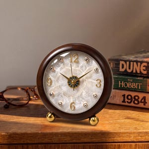 May include: A round analog table clock with a dark brown wooden frame and two small gold-toned spherical feet. The clock face features a white marbled stone texture, gold-colored numerals at 3, 6, 9, and 12, and small embedded crystals.