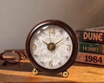 May include: A round analog table clock with a dark brown wooden frame and two small gold-toned spherical feet. The clock face features a white marbled stone texture, gold-colored numerals at 3, 6, 9, and 12, and small embedded crystals.