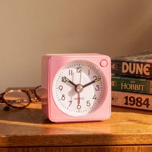 Puede incluir: Un despertador analógico cuadrado de color rosa claro con esfera blanca, números negros y manecillas de hora y minuto negras con detalles amarillos. El reloj tiene un segundero rojo y la palabra QUARTZ impresa en negro en la parte inferior.