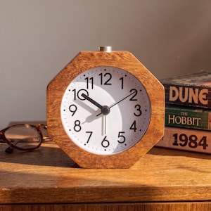 May include: An octagonal wooden alarm clock with a white face, black Arabic numerals, and black hour, minute, and second hands. A small silver button is located on the top centre of the wooden frame. The clock displays the time at approximately 10:30.
