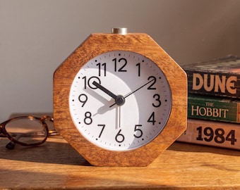 May include: An octagonal wooden alarm clock with a white face, black Arabic numerals, and black hour, minute, and second hands. A small silver button is located on the top center of the wooden frame. The clock displays the time at approximately 10:30.