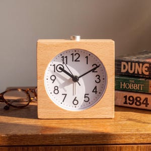 May include: A square light-colored wooden alarm clock with a white circular face, black Arabic numerals, and black hour, minute, and second hands. A small silver button is located on the top center of the wooden frame for alarm settings.