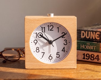 May include: A square light-colored wooden alarm clock with a white circular face, black Arabic numerals, and black hour, minute, and second hands. A small silver button is located on the top center of the wooden frame for alarm settings.