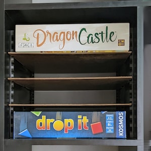 Op de afbeelding: Twee bordspellen op een plank. Het bovenste spel heet "Dragon Castle" en het onderste spel heet "Drop It".