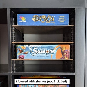 Op de afbeelding: Twee bordspellen, Dixit en Seasons, worden getoond op planken in een zwarte boekenkast. De planken zijn niet inbegrepen.