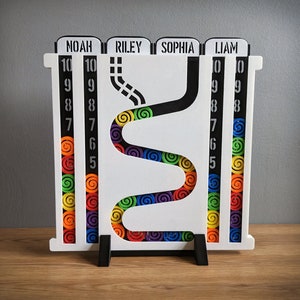 Pode incluir: Um jogo de madeira branco com os nomes "NOAH", "RILEY", "SOPHIA" e "LIAM" no topo. O jogo apresenta um caminho preto com detalhes de espirais coloridas e colunas numeradas de 5 a 10. Está sobre uma superfície de madeira.