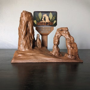 May include: A wooden model of a natural rock formation, featuring a large archway and a smaller rock spire. A sign with the text "MOAB 240" is attached to the model, with additional text reading "Dave Mori #161, 2022 Moab 240, 104:35:11".