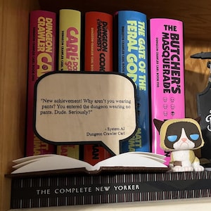 Op de afbeelding: Een boekenplank toont kleurrijke boeken, waaronder "Dungeon Crawler Carl" en "The Gate of the Feral God". Een Grumpy Cat-beeldje staat in de buurt van een bord voor "The Desperado Club". Een tekstballon citeert "Dungeon Crawler Carl".