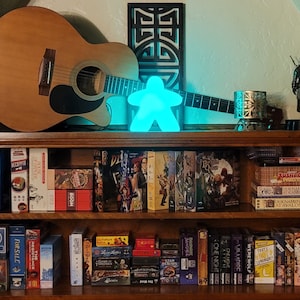Könnte beinhalten: Ein Bücherregal mit einer Vielzahl von Büchern, Brettspielen und einer türkisfarbenen Figur in Form einer Person. Das Bücherregal ist aus Holz und hat eine helle Holzoptik. Eine Gitarre lehnt an dem Bücherregal.