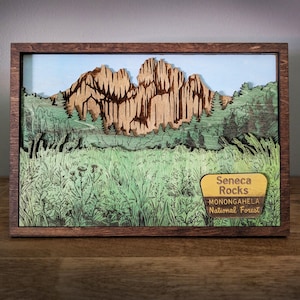 May include: A wooden framed 3D art piece depicting Seneca Rocks in the Monongahela National Forest. The artwork features a layered design with a detailed landscape of the mountains, trees, and grassy foreground. The sign reads "Seneca Rocks, Monongahela National Forest."