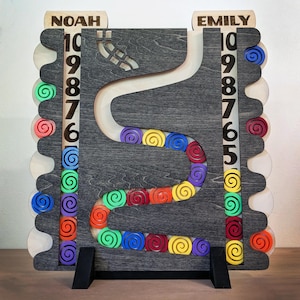 May include: A wooden chore tracker board featuring a dark gray central winding track with colorful spiral tokens. The board has two vertical columns labeled NOAH and EMILY, each with numbers 6 through 10 and side slots for storing additional tokens.