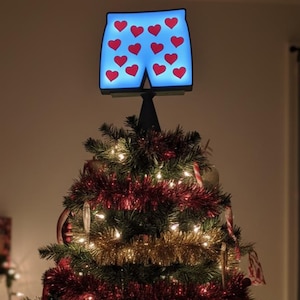 May include: A Christmas tree topped with a blue light-up decoration shaped like a pair of underwear with red hearts. The tree is decorated with red and gold tinsel, ornaments, and lights.