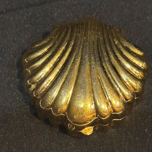 May include: A gold-toned metal compact case shaped like a scallop shell. The exterior features a series of raised, radiating ridges that mimic the natural texture of a seashell, with a polished, reflective surface and a hinged closure at the base.