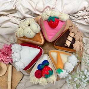 May include: A collection of six felt cake slices arranged in a circle, featuring various toppings including a red strawberry, blue berries, a carrot, and cream dollops. The slices are coloured in shades of pink, brown, orange, white, and tan.