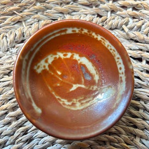 May include: A small, round ceramic bowl featuring a deep terracotta orange glaze with a prominent, lighter cream-coloured leaf-shaped pattern imprinted in the centre. The bowl has a smooth, glossy finish and a circular rim with a subtle colour variation.