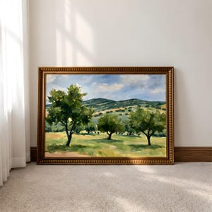 May include: A landscape painting in a rectangular gold frame featuring three green trees with yellow fruit in a grassy field. The background shows rolling hills under a blue sky with white clouds. The artwork uses visible brushstrokes.