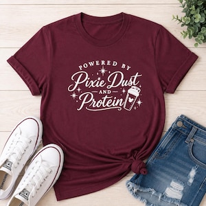 May include: A maroon short-sleeve t-shirt featuring white text that reads Powered By Pixie Dust And Protein, accompanied by a graphic of a shaker bottle with a small mouse ear icon and several starburst accents, paired with white sneakers and denim shorts.