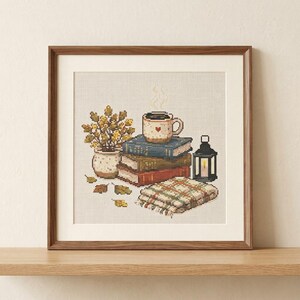 May include: A framed cross-stitch embroidery featuring a stack of three books topped with a steaming mug with a heart icon. Beside the books are a potted plant with yellow leaves, scattered autumn leaves, a black lantern, and a plaid blanket.