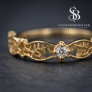 May include: A gold-toned ring featuring an intricate, openwork filigree band with a repeating leaf-like pattern and a single, small, clear round-cut gemstone set in the center. The text STONEBORNSILVER 925 STERLING SILVER is visible in the upper right.