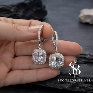 May include: A pair of sterling silver drop earrings featuring cushion-cut clear stones surrounded by halos of smaller pavé-set stones. The hinged hoops are also accented with small clear stones. The text STONEBORNSILVER 925 STERLING SILVER is visible.