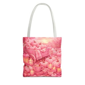 Peut inclure: Un sac fourre-tout orné d'une illustration dans les tons roses représentant une charrette en bois remplie de citrouilles. La scène comprend des lanternes lumineuses, une tasse de chocolat chaud et des citrouilles éparpillées au sol.