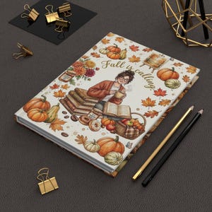 May include: A rectangular notebook featuring a white cover with a colourful illustration of a person reading among pumpkins, autumn leaves, and books. The text "Fall is calling" is printed in cursive script. The notebook is accompanied by two pencils.