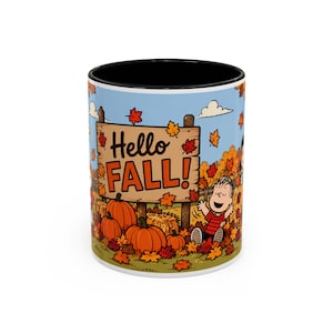 Könnte beinhalten: Ein Keramikbecher mit schwarzem Innenraum und Rand, der eine bunte Illustration einer Zeichentrickfigur zeigt, die zwischen orangefarbenen Kürbissen und Herbstlaub sitzt. Ein Holzschild im Hintergrund zeigt den Text Hello FALL in Schwarz und Orange.