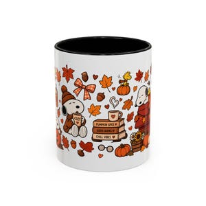 Peut inclure: Un mug en céramique blanc avec un intérieur et un bord noirs, orné d'un motif automnal coloré représentant Snoopy, Woodstock, des citrouilles, des feuilles d'érable orange, des glands et une pile de livres marqués Pumpkin Spice, Good Books, Chill Vibes.
